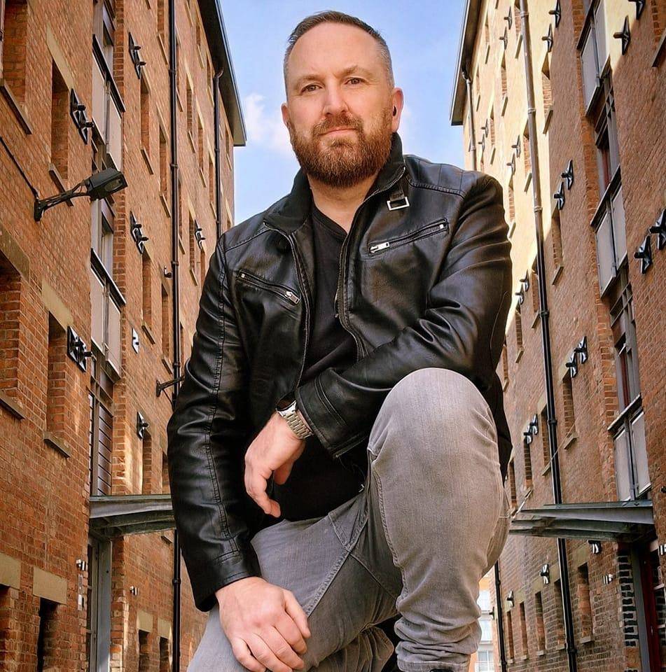 A man in a leather jacket crouching down in an alleyway.